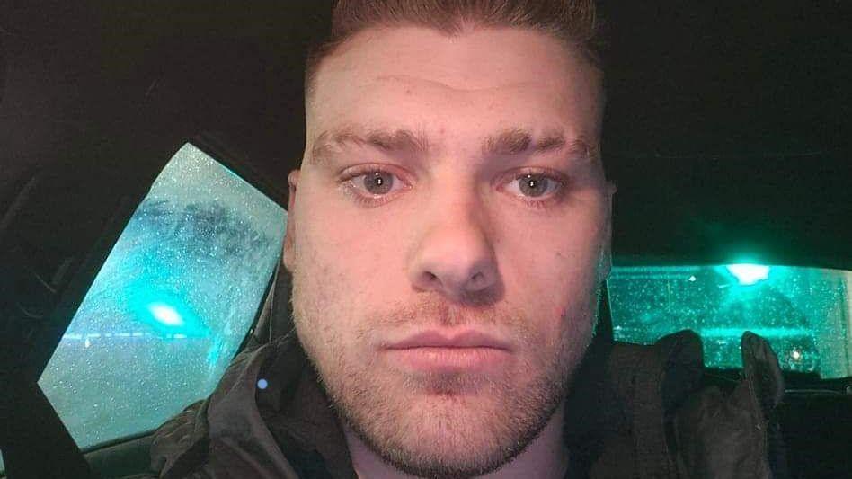 Brandon Rainey who has short brown hair and brown stubble looks at the camera. The photo is a selfie in a car - greenish light can be seen through the car windows.