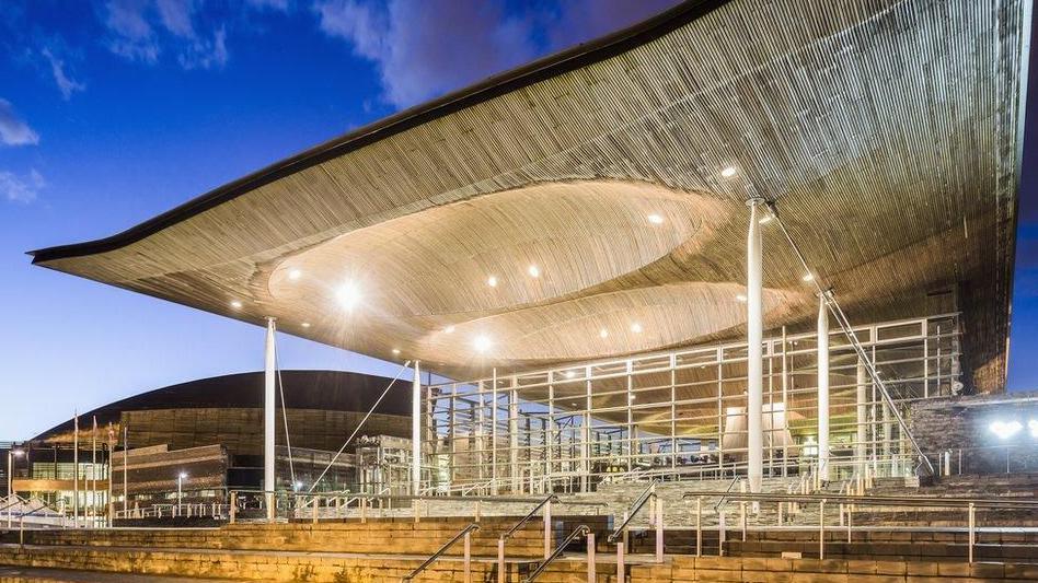 An exterior picture of the front of the Senedd building with bright lights fitted into its large roof shining down on the steps below. The Wales Millenium centre is in the background on the left hand side of the picture. 