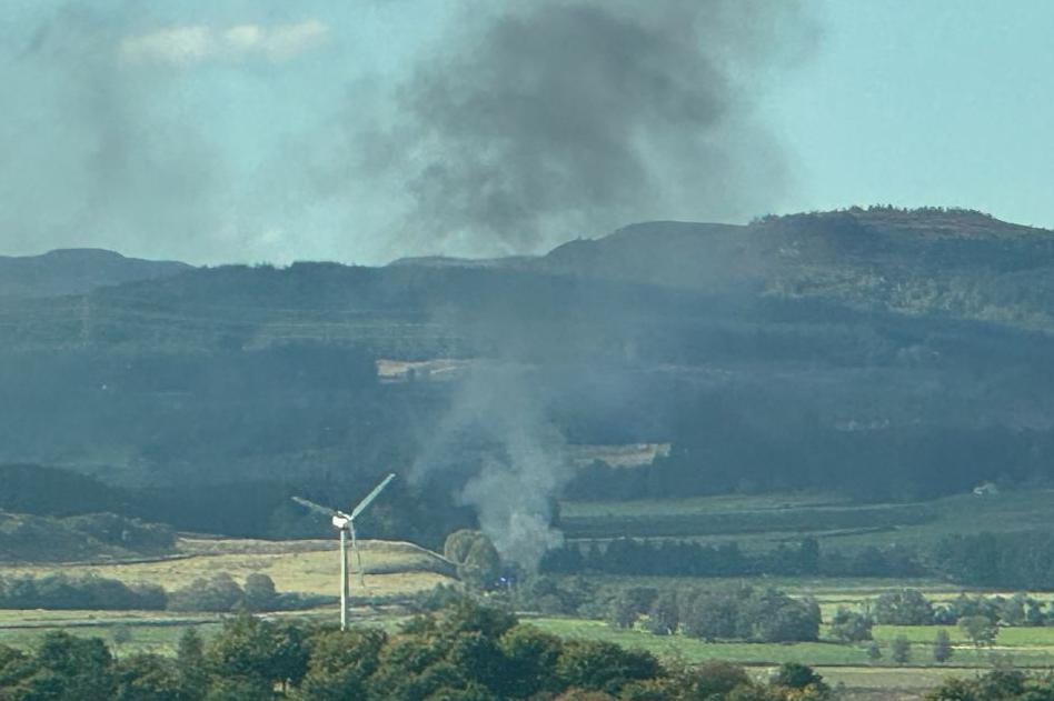 Smoke rises from the crash site. There is a wind turbine in the foreground. 