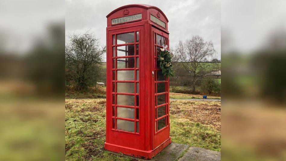 A disused telephone box in the middle of a freshly cut field. There are grey slate slabs beside it, and cut grass strewn all over the ground. It is a wet overcast day and the sky is grey. The Perspex panels on the phone box are clouded and the panel at the top which reads 'Telephone' is barely legible. A Christmas wreath has been placed on the door with a white bow on it.