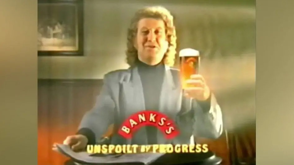 Noddy Holder wears a black top and suit jacket over the top while holding a pint of Banks's and reading a paper in a TV advert in the 80s. The brewery's advertising slogan "Unspoilt by Progress" appears on screen.