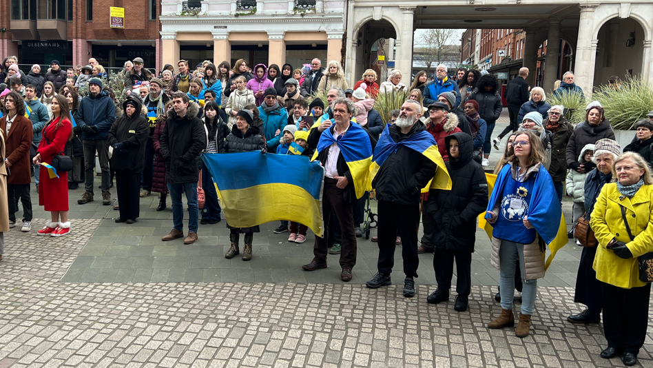 People standing in a town centre, many holding the Ukrainian flag. They appear to be listening to a speaker, who is out of shot. 