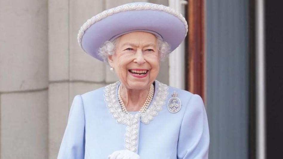 The late Queen Elizabeth II smiles broadly as she stands outside, wearing a pale blue hat and jacket; her white gloves can just be seen and she has several strands of pearls around her neck