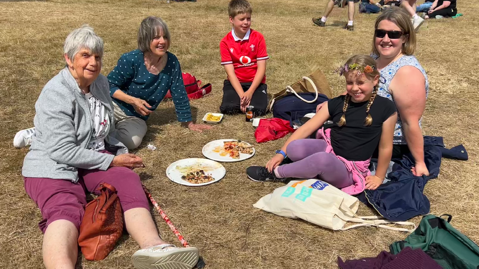 Laura is sat down on the grass with her family in a semi circle with two pizzas in the middle. Laura is on the right, just behind her daughter. Her son, mother and mother in law are also sat smiling. 