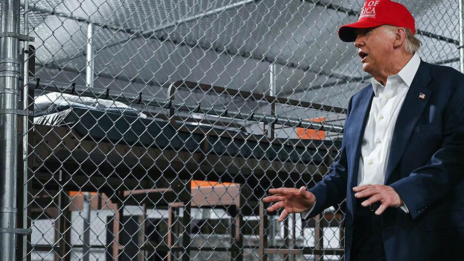 Donald Trump standing in front of a fenced off bedspace at Alligator Alcatraz wearing a red hat on 1 July 2025.