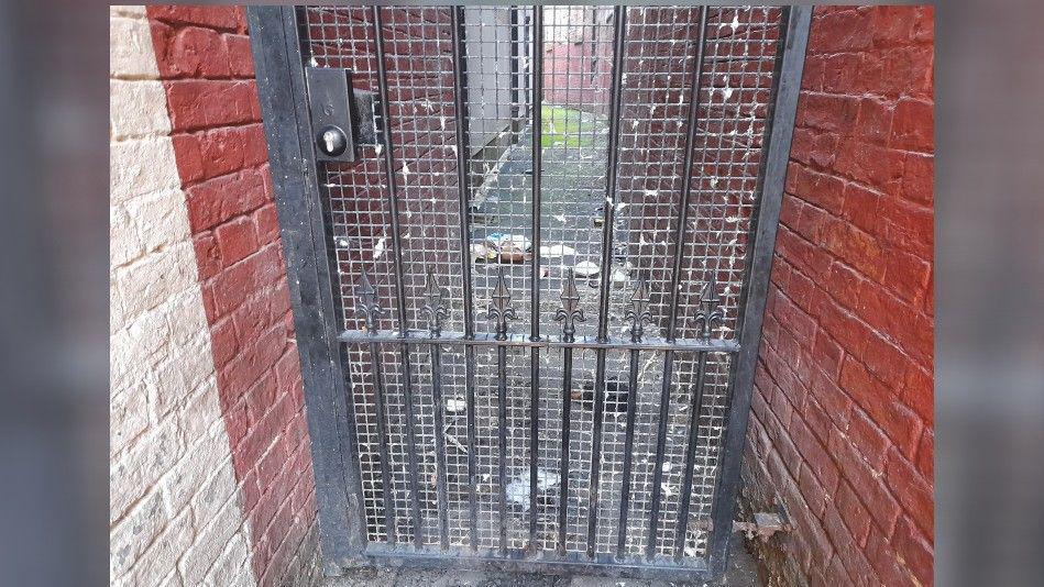 Close-up photo of a black gate which you can see through into a narrow passageway. Various litter can be seen on the other side of the gate.