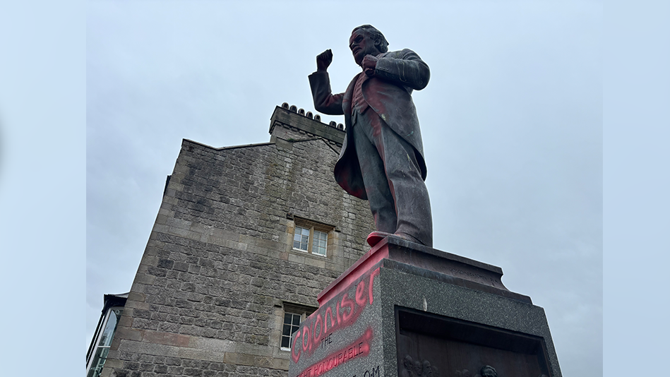 A side view of the red graffiti with the words 'coloniser' painted on the David Lloyd George statue. There is a building behind the statue with two windows. 