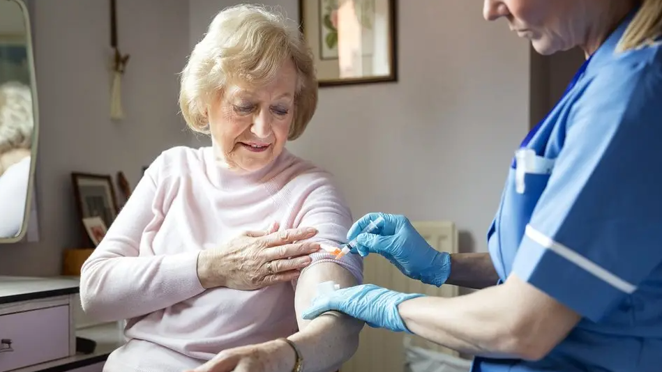 An elderly lady has her sleeves rolled up as she gets a jab. She has short blonde hair and is wearing a pink jumper. A female nurse in uniform is standing in the right of the image and has gloves on. Her face is not fully seen.