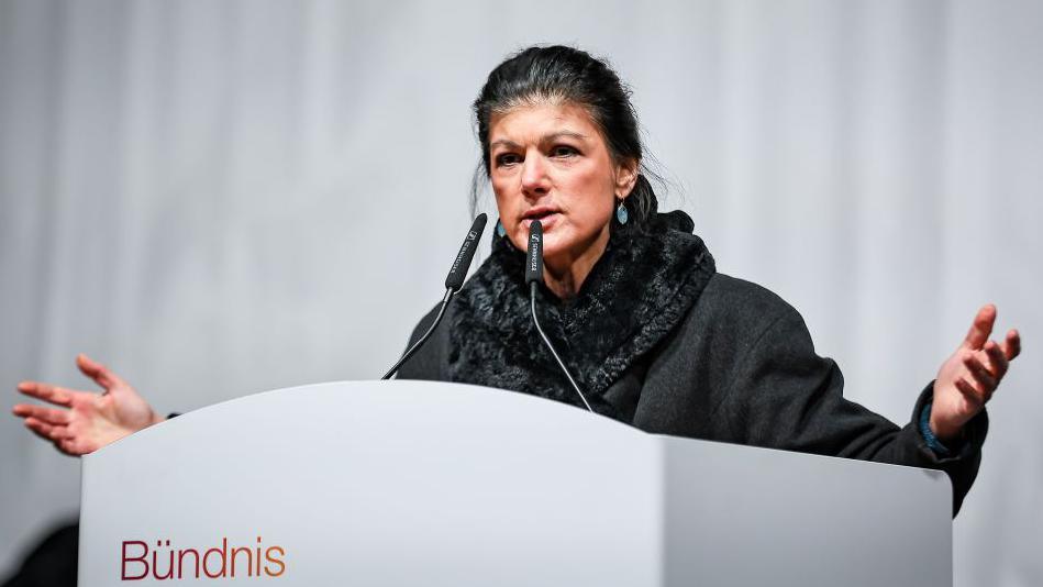 Standing on a stage, Wagenknecht launches her campaign behind a lectern wearing a large grey scarf