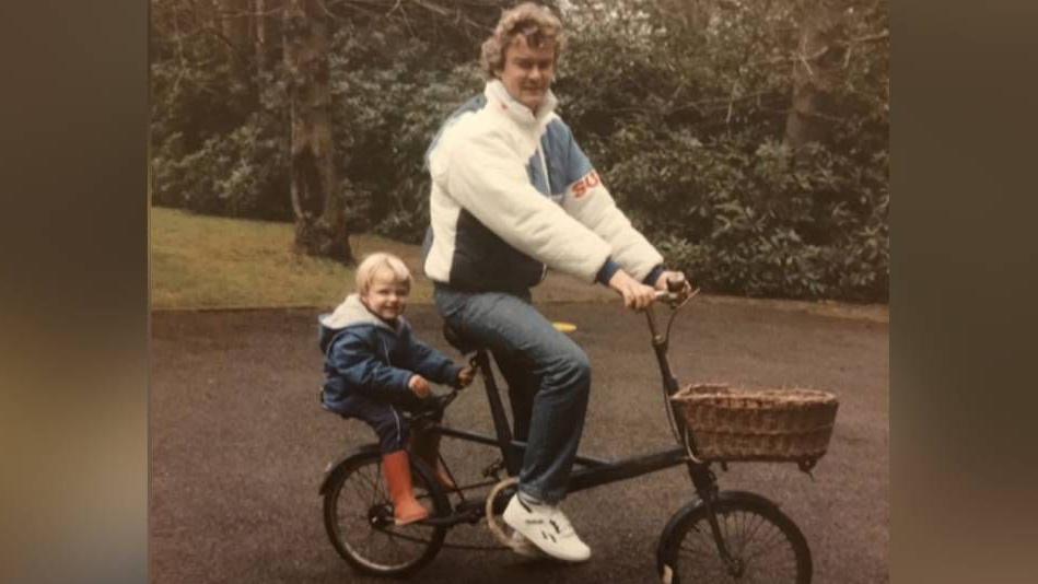 A little boy, wearing a hooded blue coat and orange wellies, sits at the back of a tandem bike with a basket at the front, ridden by his father, who is wearing a white sports jacket, blue jeans and white trainers. The little boy is grinning at the camera, but his father is not smiling.
