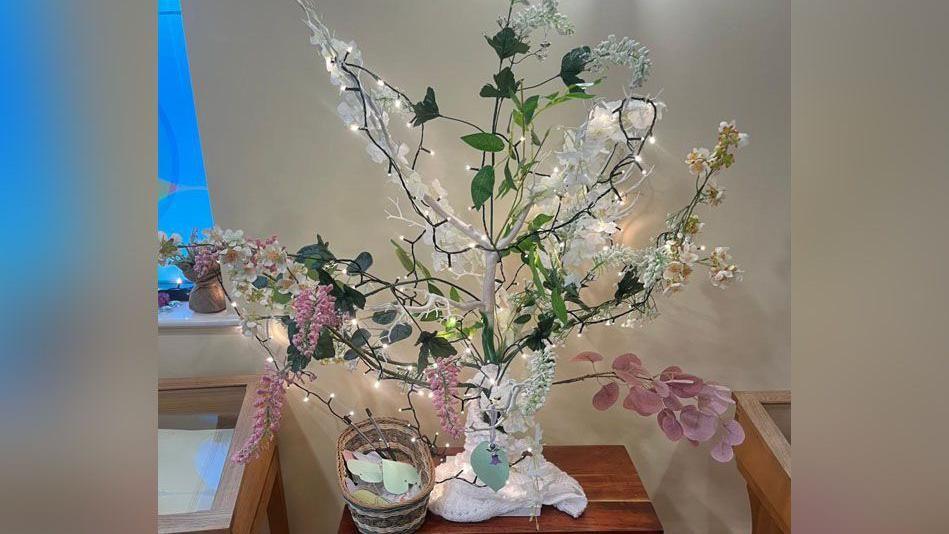 A small remembrance tree covered in fairy lights and flowers. It is indoors and perched on a small stool with a basket with heart shaped paper petals, ready for people to write on