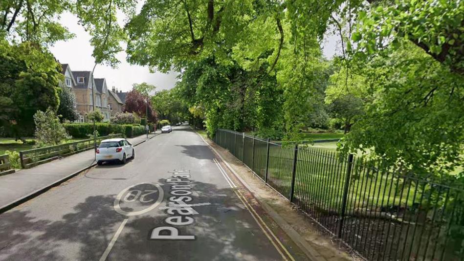 A road surround a park. On the right of the image is a large oak tree and a wrought iron fencing