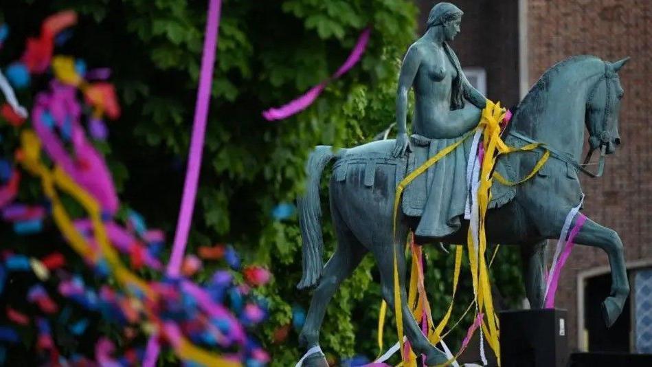 The green statue of Godiva in Coventry, with a number of colourful paper streamers draped over it. There are trees and a building in the background.