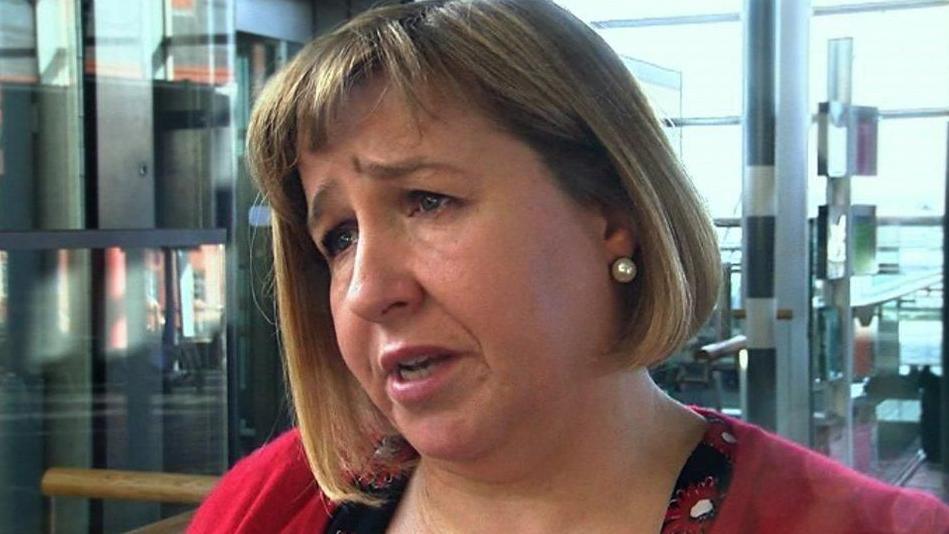 Lynne Neagle, with a red top, in front of part of the Senedd. It is a head and shoulders shot of her.