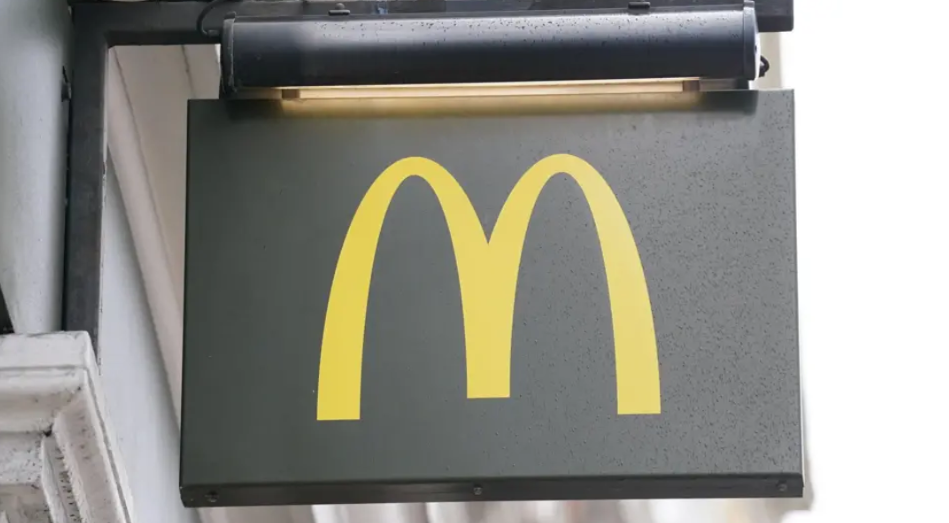 Signage for a branch of McDonald's. It is a dark grey sign with a yellow 'M' and it is it hanging on the wall of a white building.