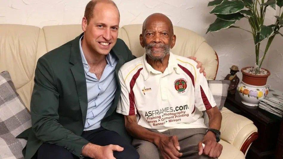 Prince William sits on a sofa with his arm round Alford Gardner, as they both smile into the camera. The Prince is wearing a green jacket and light blue buttoned shirt, while Mr Gardner is wearing a cricket top.