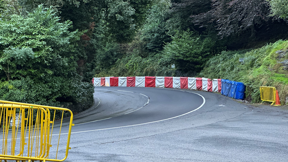 The Isle of Man TT course sweeping uphill around the bend at Glen Helen, with green trees and bushes on both sides. Red, white and blue crash barriers follow the road along the right-hand side.