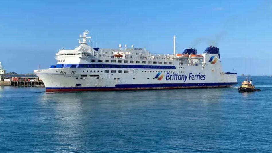 A white and blue Brittany Ferries vessel on the water with a tug boat behind it.