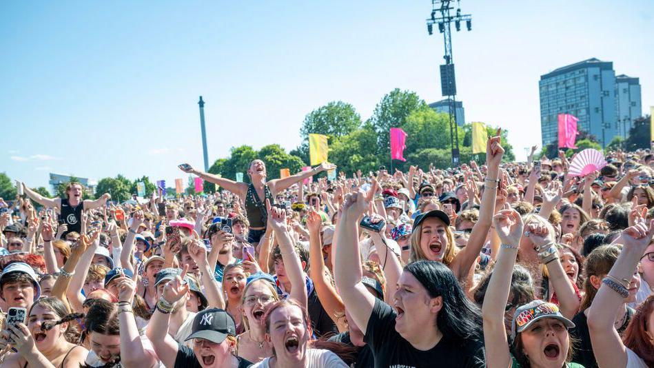 The crowd at TRNSMT from behind the barriers. People are singing with their hands in the air. It's a clear day and buildings can be seen in the distance.