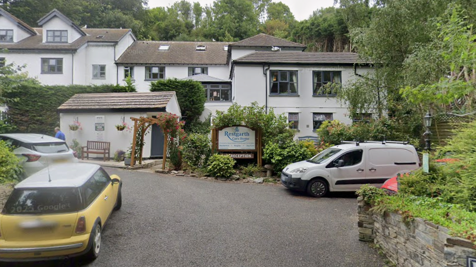 A street view image of the exterior of Restgarth Care Home. It is a small collection of white buildings with tiled rooves, nestled in between trees.