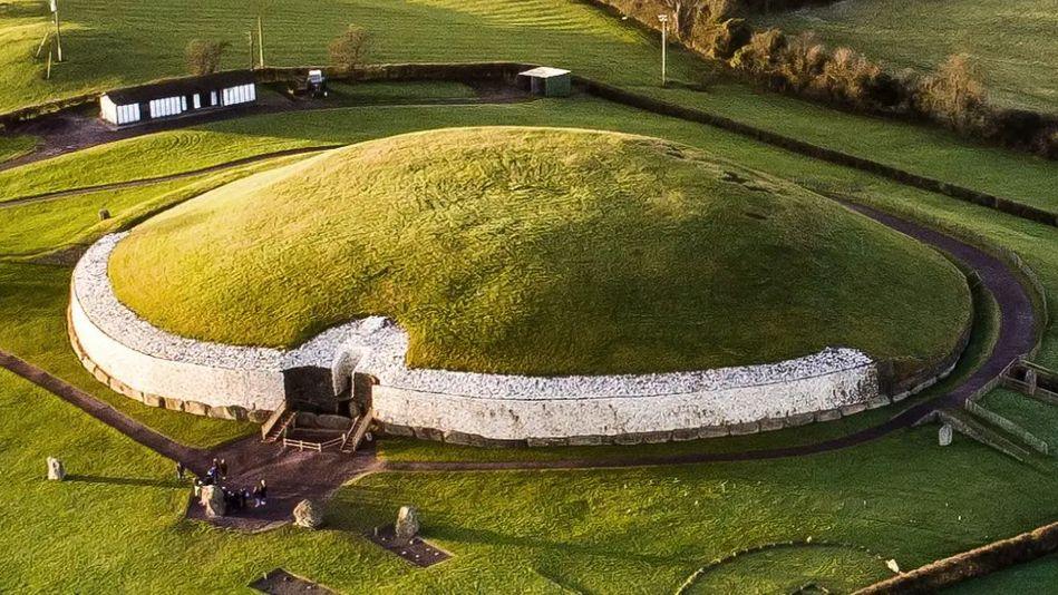 Newgrange - a mound passage tomb. It has a white stone frontage capped with a grass-covered dome. Grass surrounds it.
