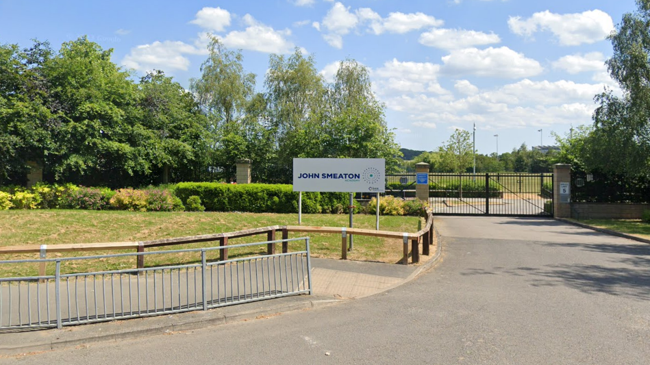 A sign outside the school indicating its name. The school gates are black and closed. Lots of trees and other street furniture can be seen.