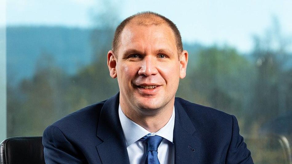 Martin Pibworth, the new chief executive of SSE, wears a navy suit and blue tie while sitting at a desk, smiling, with large windows and a blurred outdoor background behind him.