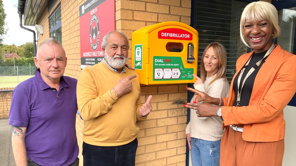 Roy Tucker in a purple T-shirt and trousers, Kolin Bhogal in a shirt, yellow jumper and black trousers, Louise Astley in a beige jumper and jeans and Celia Hibbert wearing an orange trouser suit, stand at the entrance to the club next to the yellow defibrillator attached to the wall of the building.