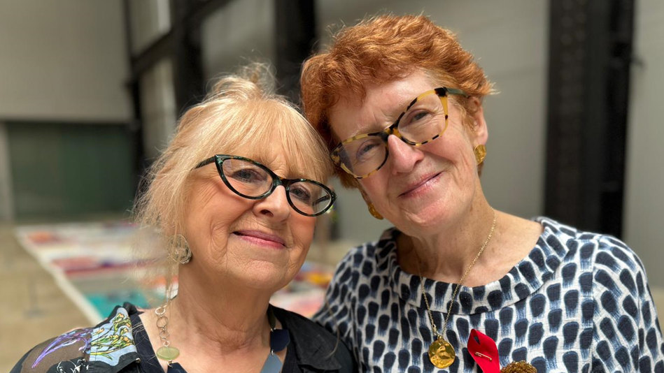Cathy and her friend Grace smile at the camera. They are both older women wearing glasses and brightly coloured tops and jewellery. They are standing in a hall with parts of the quilt on the floor behind them.