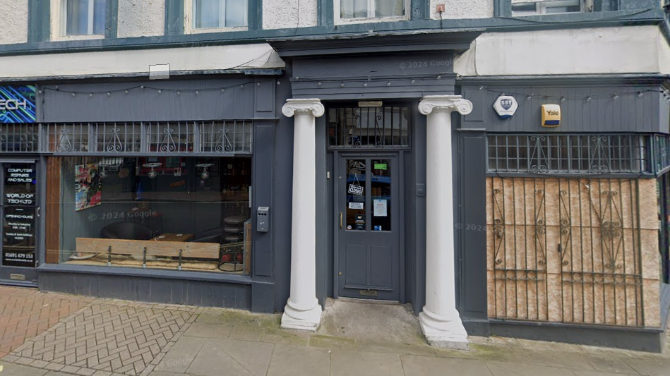 The front of a shop that is made from wood and painted dark grey. On either side of the doorway, there are two white pillars. Above the large shop window there is a small canopy.