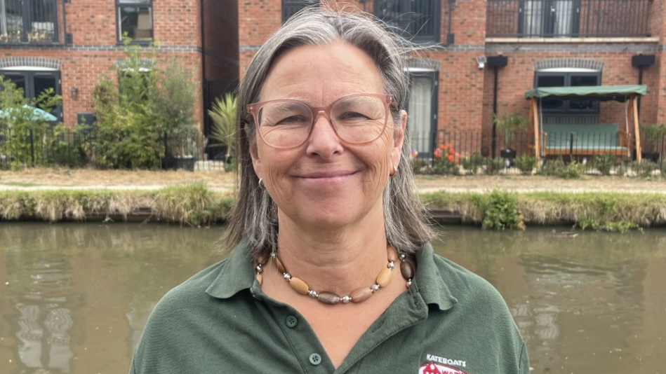 A woman with shoulder length grey hair, large glasses and a green polo shirt standing in front of an area of green water and a tall brick building with black metal fencing in front of it