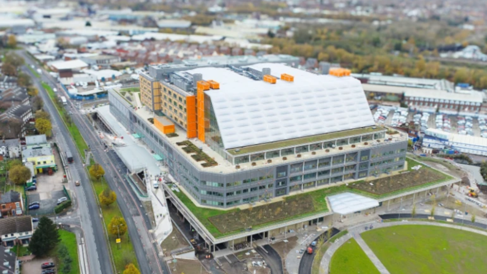 Aerial view of a very large rectangular building, with grey floors at the bottom, and a white roof with orange elements to the side and top.