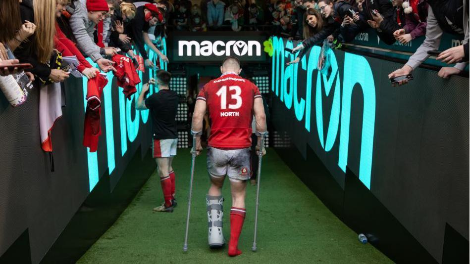 A view from behind of George North walking down the players' tunnel on crutches and with left leg in a protective boot 