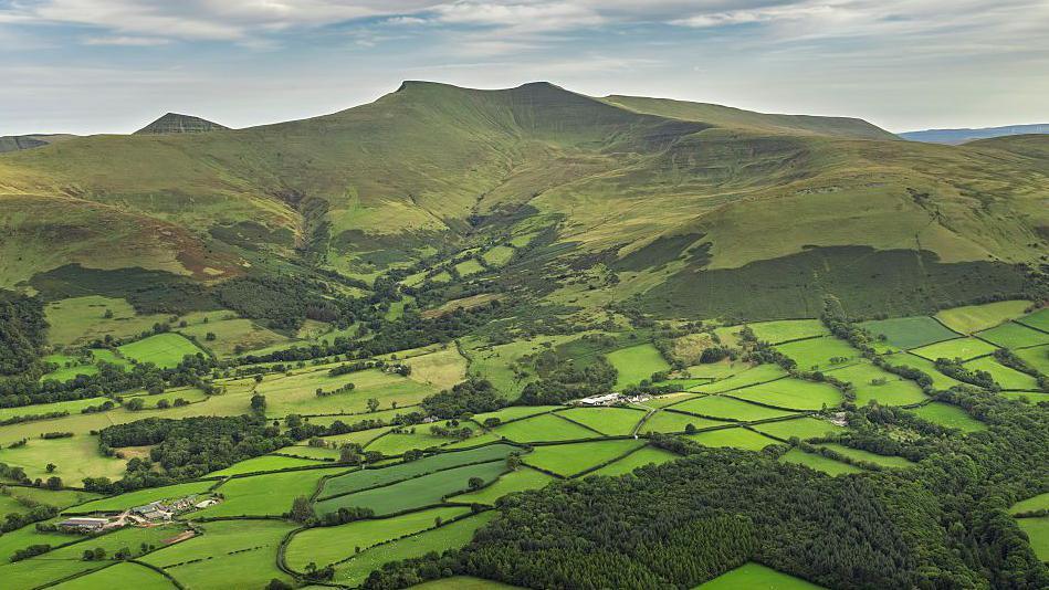 An aerial view captures of the Brecon Beacons, surrounded by green forests and plains.