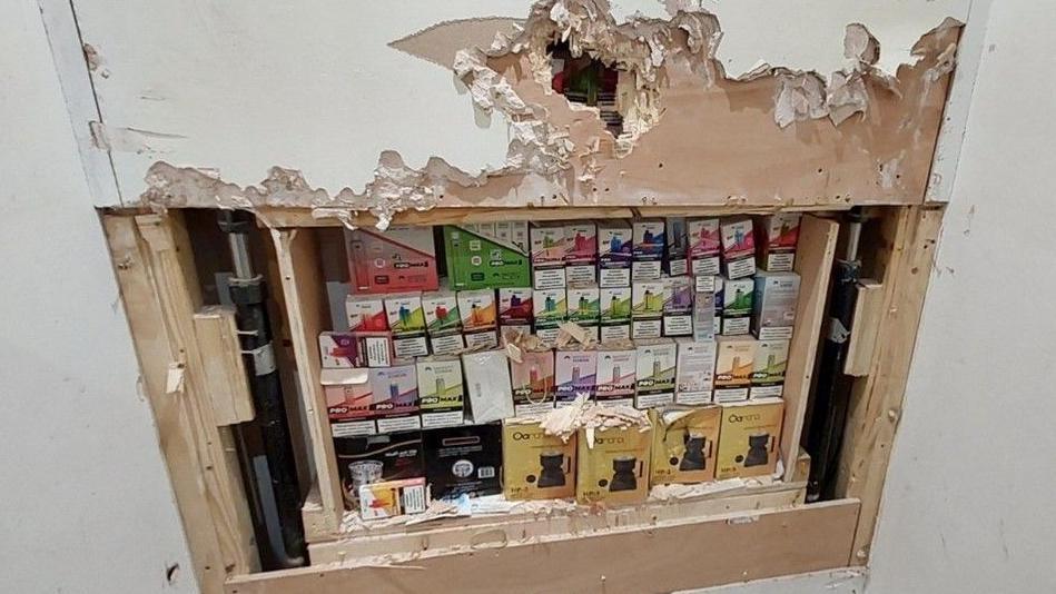 A large collection of illegal vapes in colourful boxes stashed behind a wall, which has been peeled back by officers.