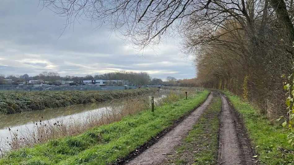 A mud track runs alongside a rive. It is shelted by a high hedgerow, has no lights and looks gloomy.