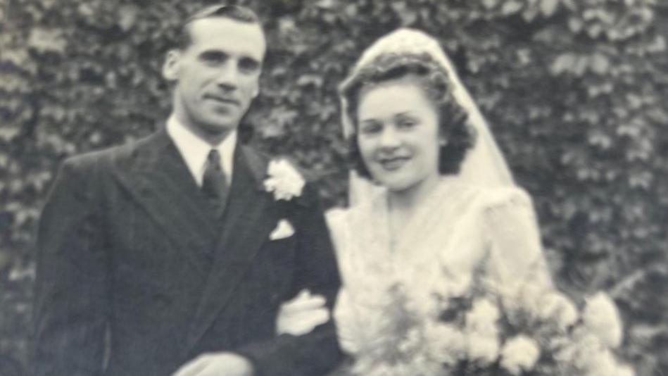 Phyliss Marshall's wedding photo taken in 1948, she is pictured wearing her white dress with a large collar and veil holding a bouquet of flowers. Both are arm in arm.