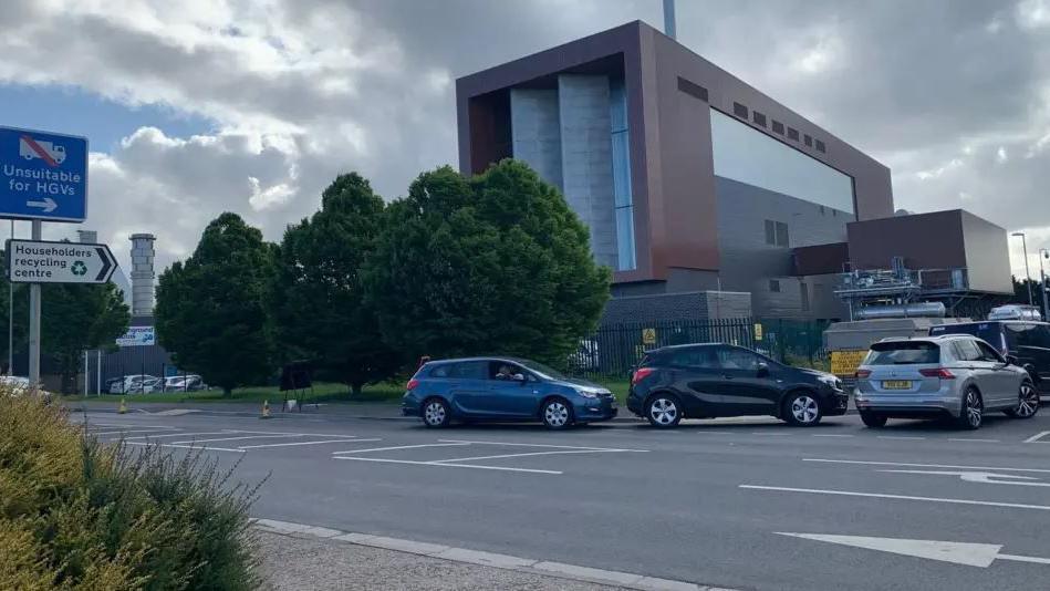 A large rectangular building with a queue of cars on the road outside 