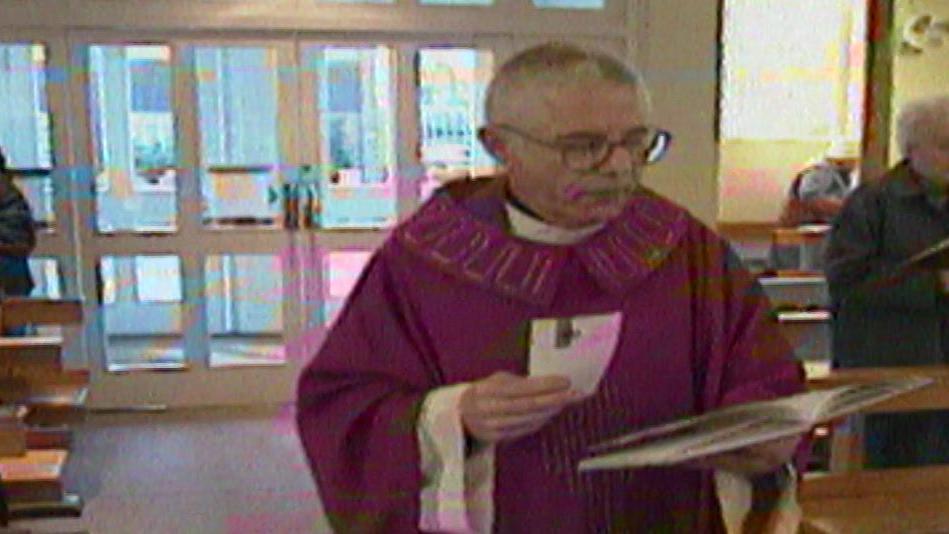 Archive Look East footage of Dennis Finbow wearing a purple liturgical robe, standing near the entrance of a church. He holds a booklet and a piece of paper, with pews and other individuals praying in the background.