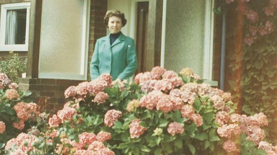 A woman wearing a blue coat stands outside a house. Pink flowers can be seen in front of her.