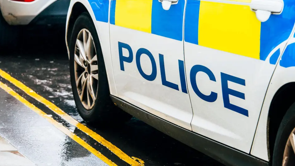 The side of a police car, with "police" emblazoned on it in blue on a white background, parked on the side of a wet road.