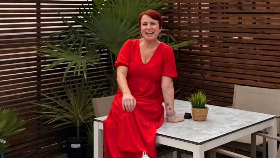 Clare Kendall is wearing a red dress and has red hair is sitting on a garden table smiling at the camera.