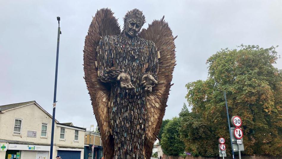 The Knife Angel statue rises into the sky. It is composed of thousands of knives. In the background are trees and a row of shops. 