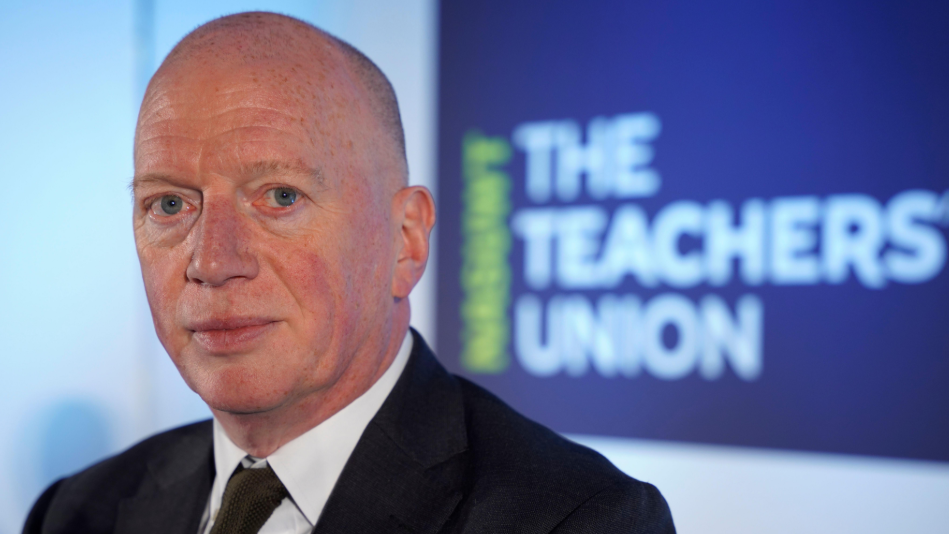 Matt Wrack is wearing a suit and is sitting in front of a poster which says the teachers union