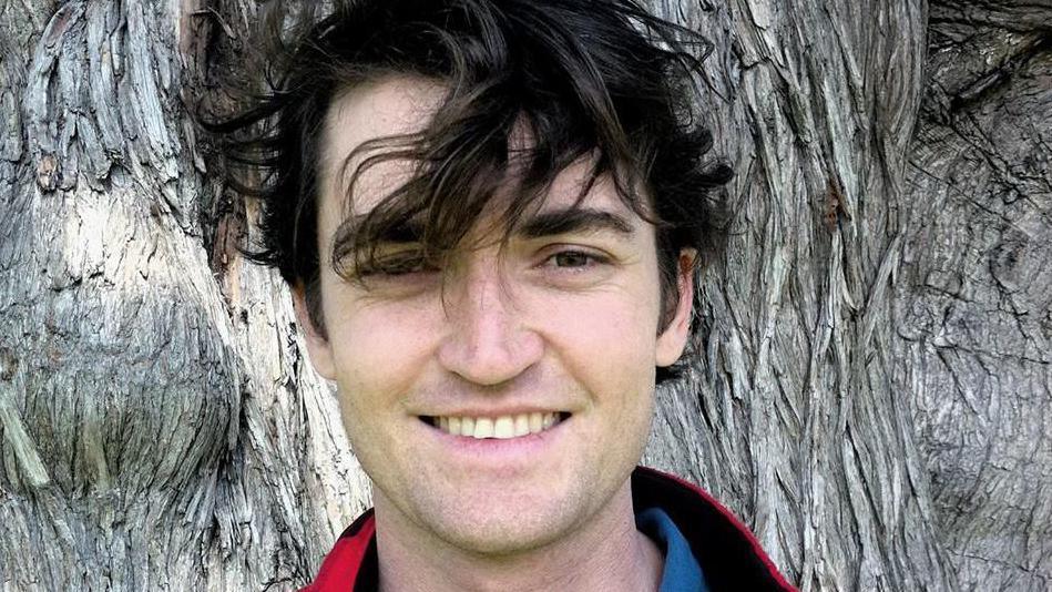 A smiling Ross Ulbricht is pictured in front of a large tree trunk, his hair blowing in the wind. 