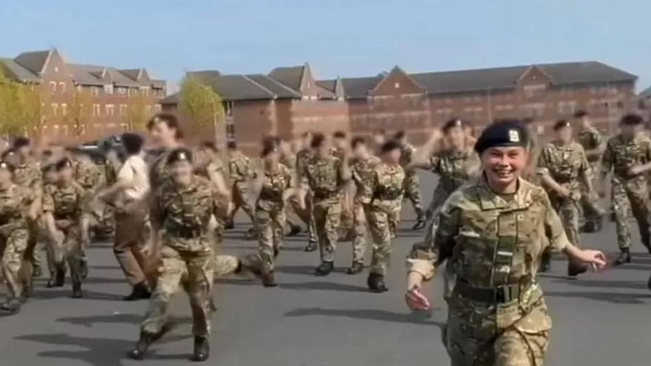 A group of soldiers in camouflage uniforms and berets running on a parade ground. A young girl in the same uniform is running towards the camera.  