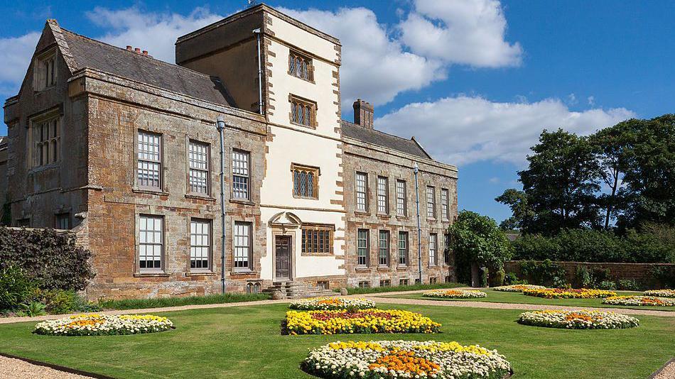 A shot of a four storey stately home with red and white bricks and a well kept lawn with yellow, white and orange flower arranged in a circular pattern.