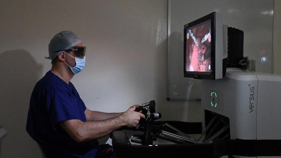A surgeon is sat with his hands on a console, which looks like robotic arms that are attached to the Versius. He is looking at a screen. He is wearing dark blue scrubs, a blue mask and black goggles.