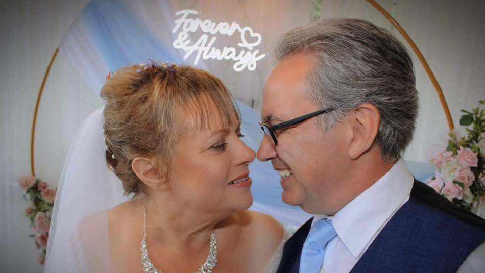 Newlyweds Nikki Flux-Edmonds and Patrick Hedges standing next to each other, seemingly about to kiss each other. They are standing underneath a sign that says "forever and always".