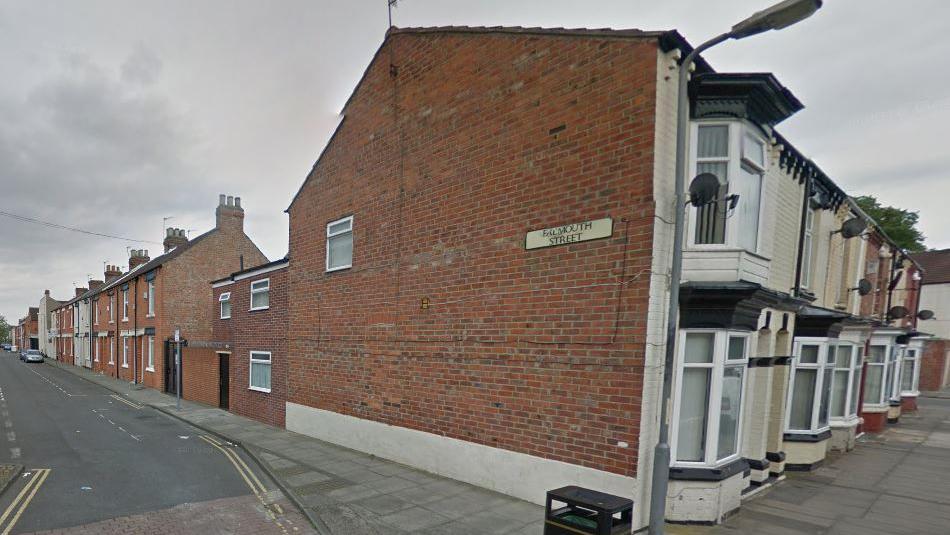 Falmouth Street in Middlesbrough. The street sign is attached to the side of a terraced house on an adjacent building. A row of red-brick terrace houses line Falmouth Street. There are parking bays in the road but few cars.